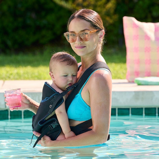 Woman holding a baby in a sling by a pool with a pink chair and greenery in the background